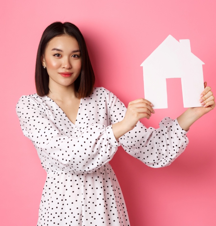Real estate. Beautiful asian woman demonstrating paper house model, looking at camera confident, advertising home for sale, standing over pink background.