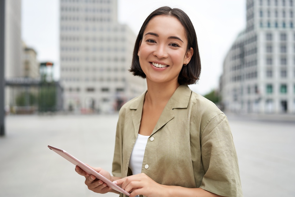 Young smiling asia woman with tablet, standing on street in daylight.